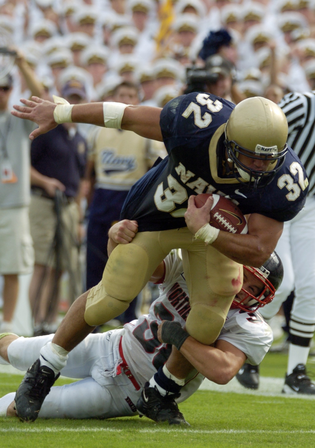 Zwei Fußballspieler auf dem Feld, einer im Sportdress und Helm, der von einem anderen am Boden liegenden Spieler getackelt wird, mit anderen Spielern und Zuschauern im Hintergrund.