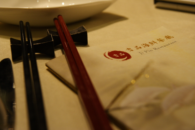 Ein Tisch mit einer Speisekarte, die 'Ji Pin Restaurant' auf Mandarin und Englisch lautet, sowie zwei Paar Stäbchen (eins rot, eins schwarz) und eine weiße Schüssel vor den Stäbchen.