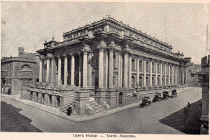 Ein altes Schwarz-Weiß-Foto des Teatro Massimo Opernhauses in Rom, Italien, mit einem großen Gebäude mit Säulen, Treppen und Bögen, sowie einigen Menschen und Karren auf der Straße davor und Text am unteren Rand des Bildes.