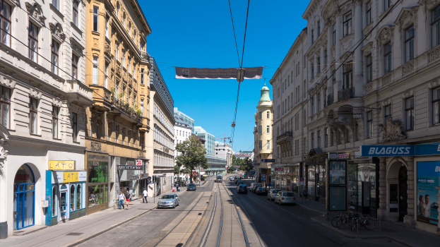 Eine Stadtstraße mit einer Tram auf den Schienen, Fahrzeugen auf der Straße, Fußgängern auf dem Gehweg, parkende Fahrräder auf der rechten Seite, Gebäude mit Fenstern auf beiden Seiten und Bäume unter einem klaren blauen Himmel im Hintergrund.