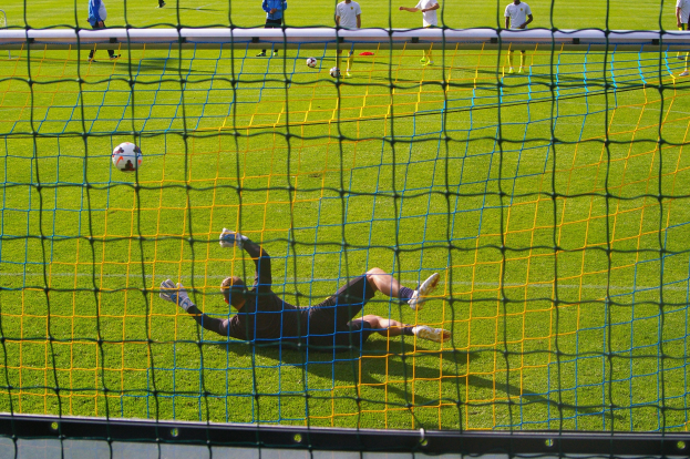 Ein Fußballtorhüter beim Abwehren eines Schusses vor einem Tor mit Netz im Vordergrund und einigen Menschen im Hintergrund, sowie mehreren Fußballen auf dem Boden.
