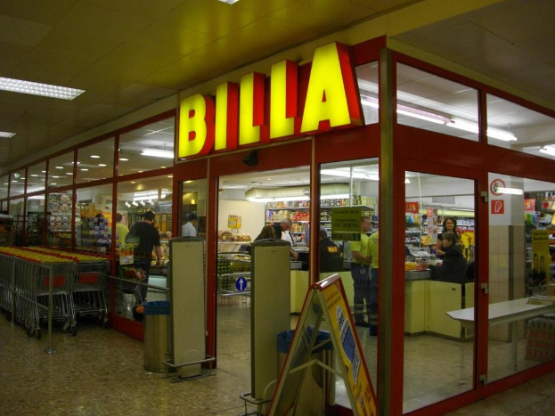Ein Markt mit einem Eingang in der Mitte, Mülltonnen daneben, eine Reihe von Einkaufswagen auf der linken Seite, Deckenbeleuchtung und einige Menschen, die drinnen gehen.