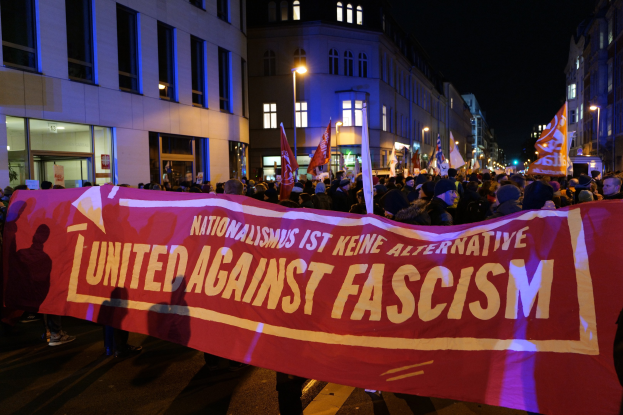 Eine Gruppe von Menschen marschiert mit einem Banner, auf dem 'Einheit gegen Faschismus' steht, eine Straße entlang, mit Gebäuden, Laternen und Schildern an der Straße und einem sichtbaren Himmel.