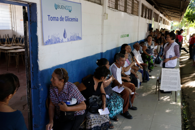 Eine Gruppe von Menschen sitzt auf Bänken vor einem Gebäude mit einer Texttafel und einer Tür, die in einer Warteschlange zum Abstimmen an einer Wahllokal in Guadalajara, Mexiko, mit Bäumen im Hintergrund warten.