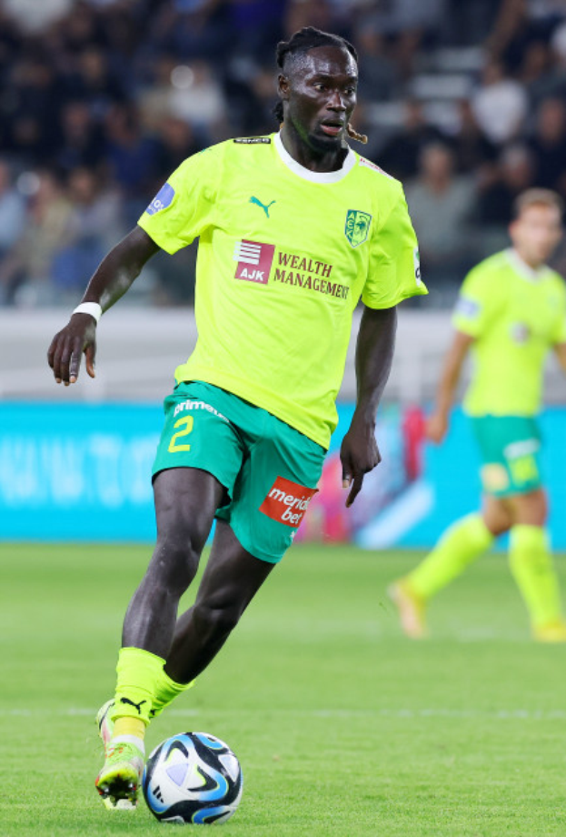 Ein Fußballspieler in gelbem Trikot und grünen Shorts ist im Einsatz auf dem Feld, mit einer blauen Tafel und unscharfen Figuren im Hintergrund.