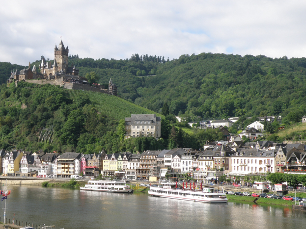 Ein malerischer Blick auf den Rhein in Deutschland mit einer Burg auf einem Hügel, Booten auf dem Fluss, Fahrzeugen auf einer nahen Straße und einem bewölkten Himmel.