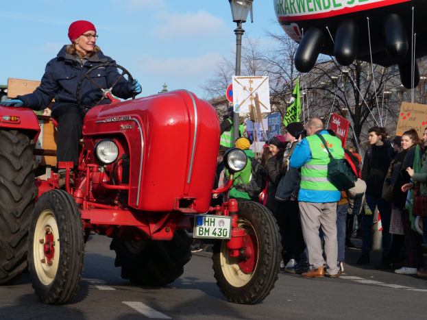 Eine Frau fährt einen roten Traktor eine Straße entlang, umgeben von einer Menge mit Schildern und Transparenten, mit Laternenmasten, Bäumen, Gebäuden und einem klaren blauen Himmel im Hintergrund.