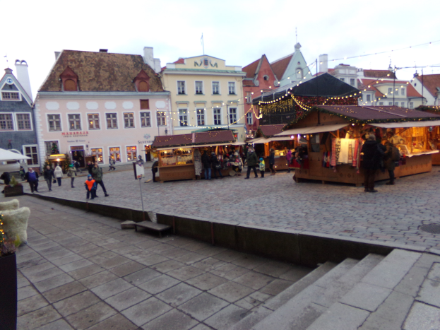 Ein belebter Weihnachtsmarkt in Tallinn, Estland mit Menschen um geschmückte Stände, festliche Lichter, Stufen nach oben, Topfpflanzen und Gebäuden im Hintergrund unter einem bewölkten Himmel.