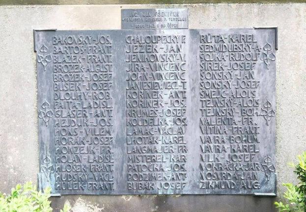 Ein Steinplakette an einer Wand mit eingraviertem Text, umgeben von Pflanzen am Boden, als Denkmal für Holocaust-Opfer.