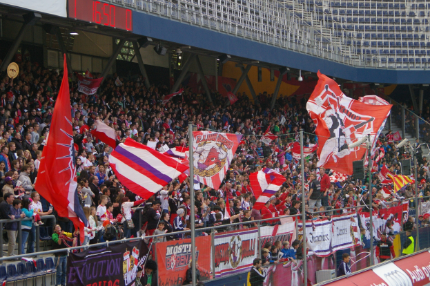 Eine große Menschenmenge in einem Stadion mit Fahnen und Transparenten, umgeben von einer Begrenzung und einem digitalen Anzeigebrett oben.