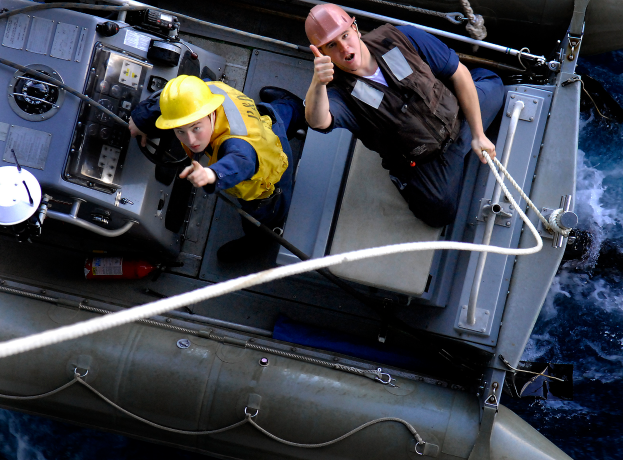 Zwei Männer in gelben Helmen und Sicherheitswesten auf einem Boot, einer hält ein Seil, scheinen eine Rettungsaktion in der Mitte eines Gewässers durchzuführen.