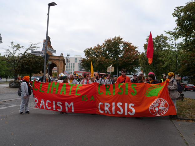 Gruppe von Menschen marschiert auf einer von Bäumen gesäumten Straße und hält ein Banner mit der Aufschrift "Klimakrise ist eine Krise" hoch, während Fahrzeuge vorbeifahren und Gebäude im Hintergrund zu sehen sind, unter einem klaren blauen Himmel.