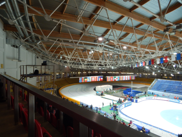 Große Indoor-Eisfläche mit zahlreichen Menschen, die Schlittschuh laufen, umgeben von Sitzplätzen, Geländern, Brettern, Beleuchtungskörpern und anderer Ausstattung, mit einer Wand im Hintergrund.