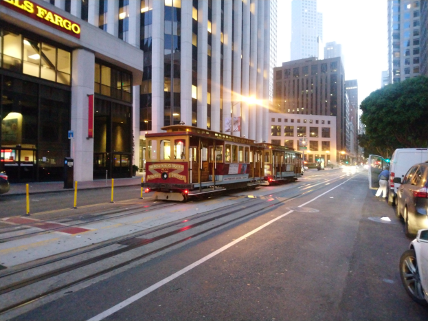 Eine Seilbahn fährt an einer städtischen Straße in San Francisco entlang, mit Fahrzeugen, einem Fußgänger und einem geparkten Van auf der Straße.
