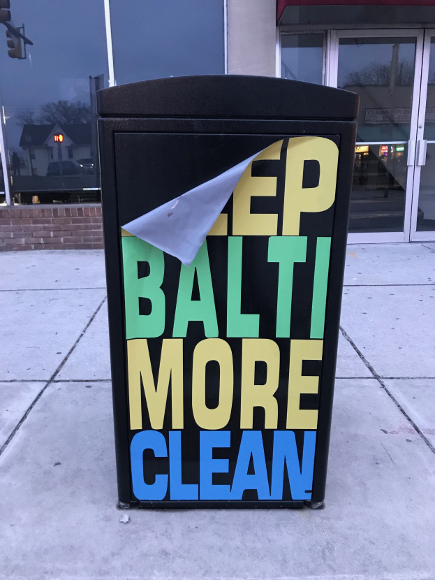 Ein schwarzer Müllcontainer mit der Aufschrift "Keep Baltimore More Clean" vor einem Gebäude mit Glastüren auf dem Bürgersteig.