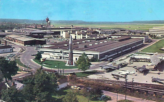 Altes Schwarz-Weiß-Foto eines Flughafens mit parkenden Flugzeugen, Gebäuden, Bäumen, Straßen, Fahrzeugen, Strommasten, Gras, Bahnschienen, Türmen, Hügeln und einem klaren Himmel mit Text am unteren Rand.