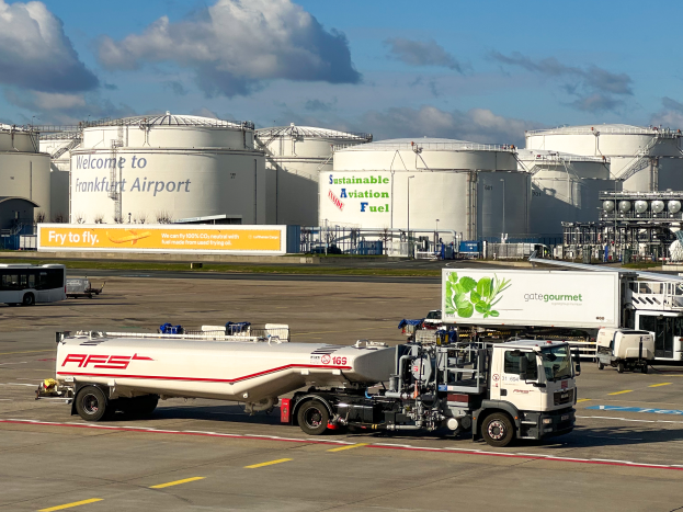 Ein großer weißer und grüner Tanklastzug parkt vor einem Gebäude am Frankfurter Flughafen. Im Hintergrund sind andere Fahrzeuge, große weiße Tanks und ein gelbes Schild unter einem klaren blauen Himmel zu sehen.