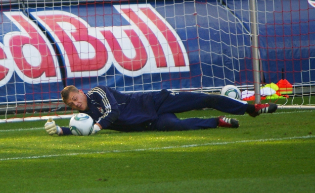 Ein Fußballspieler liegt auf dem Boden mit einem Ball vor ihm, umgeben von Gras, einem Netz, einer Fahne mit Text und anderen Gegenständen.
