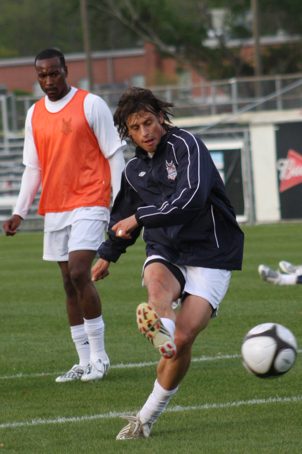Zwei Fussballspieler, einer in einer schwarzen Jacke und weissen Shorts, die einen Ball treten, und ein anderer in einer orangen Jacke hinter ihm, mit einem Zaun und Bäumen im Hintergrund.