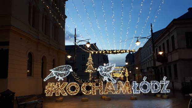 Eine belebte Stadtstraße in Krakau, Polen, geschmückt mit festlichen Weihnachtslichtern, gesäumt von Gebäuden, Straßenlaternen, Lichtern, Bänken und beleuchteten Dekorationen unter einem sichtbaren Himmel.