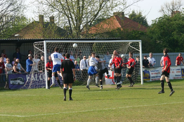 Players are playing football on a field with a goal net, while spectators stand behind it, with trees and houses in the background.