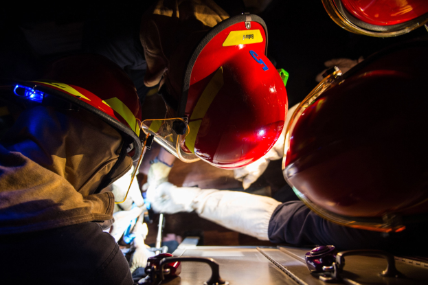 Feuerwehrleute in Helmen und Handschuhen arbeiten an einem Feuerwehrauto in einer dunklen Umgebung, mit einem Tisch voller Vorräte auf der rechten Seite.