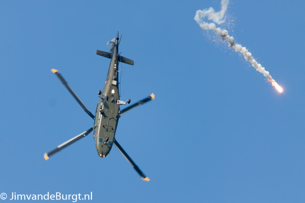 Ein militärisches Helikopterflugzeug, das eine Rakete hält, die Rauch und Feuer ausstößt, mit Text in der unteren linken Ecke.