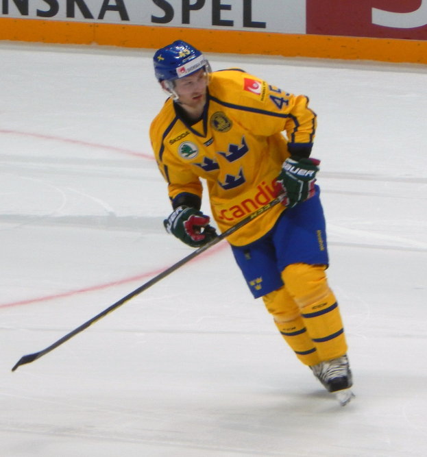 Ein Eishockeyspieler in Bewegung auf dem Eis, der einen Helm, Handschuhe und Schlittschuhe trägt und einen Hockeystock hält, mit einer Tafel im Hintergrund, auf der Text zu sehen ist.
