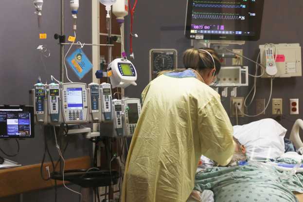 Ein Mann liegt in einem Krankenhausbett neben einem Monitor, mit einer Frau neben ihm; ein Tisch mit einem weiteren Monitor ist links und medizinische Geräte sind im Hintergrund zu sehen.