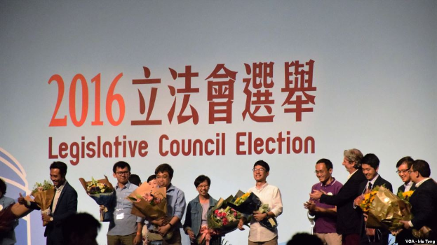 Eine Gruppe von Menschen auf einer Bühne mit Blumensträußen, mit einem Publikum darunter und einem Bildschirm mit Text und Logo, das die Wahl des Legislativrats 2016 anzeigt.