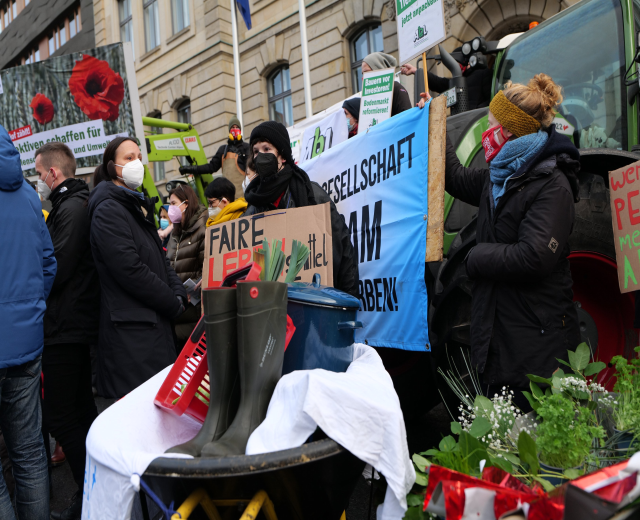 Eine Gruppe maskierter Menschen mit Protestschildern vor einem Lastwagen, mit einem Tisch voller Gegenstände, Topfpflanzen und einem Gebäude mit Flagge im Hintergrund.