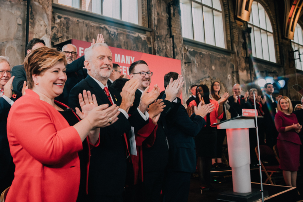 Eine Gruppe von Menschen, die vor einem Publikum applaudieren, mit einem Podium, einem Mikrofon und einer Tafel mit Text auf der rechten Seite sowie Stühlen, einem Banner, einer Wand, Fenstern und Lichtern im Hintergrund.