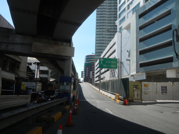 Stadtstraße mit Absperrkegeln, Fahrzeugen, einer Brücke auf der linken Seite, Gebäuden mit Namensschildern, Laternenmästen und einem Himmel im Hintergrund.