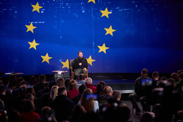 Ein Mann in einem schwarzen Kleid sitzt auf einer Bühne und hält ein Mikrofon in der Hand, mit zwei Tischen voller Gegenstände dahinter und spricht zu einem sitzenden Publikum vor einem blauen Bildschirm mit gelben Sternen.