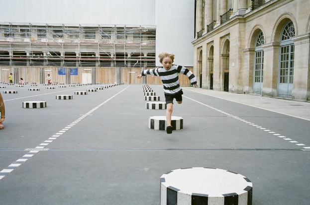 Ein Spielplatz mit vier Kindern, eines laufend auf einem schwarz-weißen Betonstufenbereich, umgeben von einem Schulgebäude.