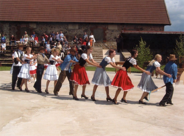 Eine Gruppe von Menschen in traditioneller bayrischer Tracht tanzt vor einem Gebäude mit Dach und Fenstern, einige tragen Hüte und halten Stöcke, während andere auf Bänken sitzen, mit grasbewachsenem Boden und Bäumen im Hintergrund unter einem bewölkten Himmel.
