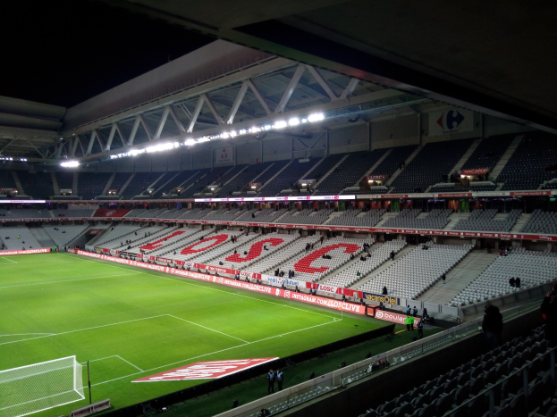 Großes Stadion mit einem grünen Fußballfeld, einem Netz auf der linken Seite, einigen Zuschauern auf den Tribünen und heller Deckenbeleuchtung vor dunklem Hintergrund.