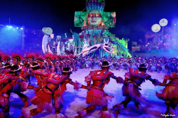 Eine Gruppe von Menschen in bunten Kostümen tanzt auf einer Bühne vor einem sitzenden Publikum, mit Lichtern, Stangen, Ballons und einem Bildschirm, der eine Person im Hintergrund zeigt, unter einem sichtbaren Himmel, bei der Rio-Karneval 2014.
