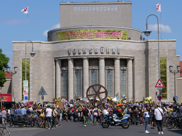 Eine große Gruppe von Menschen versammelte sich vor einem Gebäude mit Säulen und Text, umgeben von Straßenmästen, Lichtern, Schildern, Motorradfahrern, Kraftfahrzeugen, Bäumen und einem bewölktem Himmel während einer Demonstration in Berlin, Deutschland.