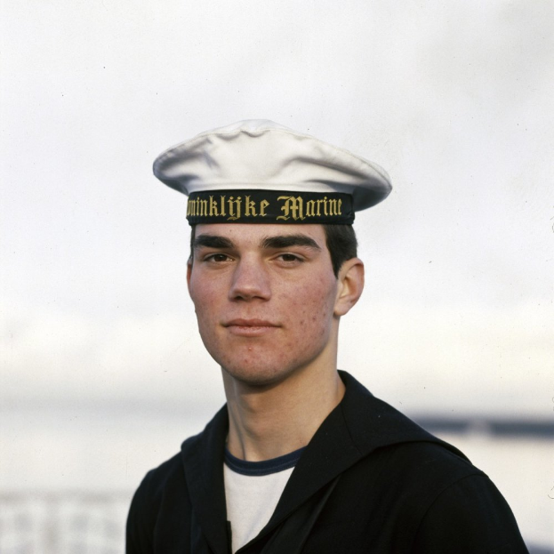 Ein Mann in einer Matrosenuniform, der eine schwarze Robe und eine weiße Mütze mit Text trägt, steht auf einem Boot mit einem unscharfen, träumerischen Hintergrund.