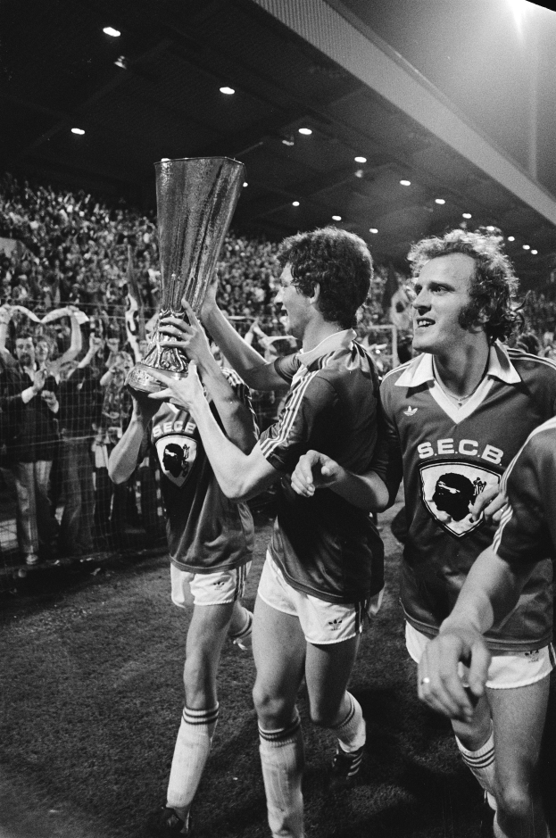 Eine Gruppe von Männern in Schwarz-Weiß-Kleidung hält einen Pokal auf einem Fußballfeld hoch, während sie ihren Sieg im FA Cup Finale feiern, umgeben von einer Menge hinter einem Zaun unter Stadionbeleuchtung.