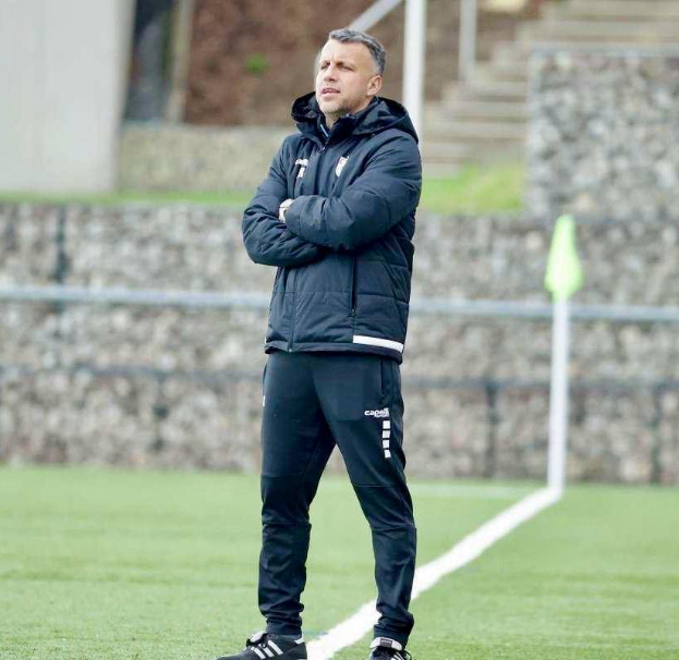 Ein Mann steht auf einem Fußballfeld mit verschränkten Armen, trägt eine schwarze Jacke und Hose und hat im Hintergrund einen Zaun, Treppe und eine Wand.