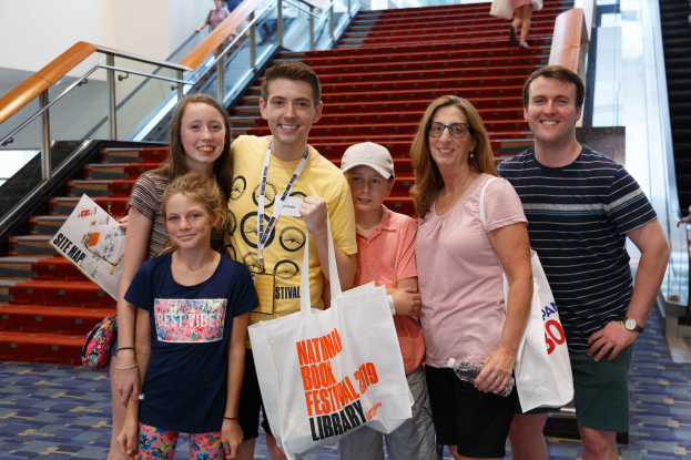 Eine Gruppe strahlender Menschen mit Taschen steht zusammen vor einer Rolltreppe auf dem Nationalen Buchfest 2019, mit Treppen, Geländern und einer Wand im Hintergrund.