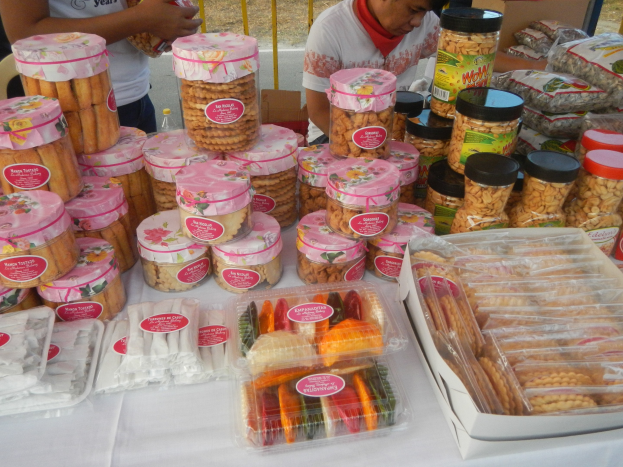 Eine Gruppe von Menschen um einen Tisch versammelt, auf dem verschiedene verpackte Lebensmittel liegen, mit einem Gel├äder und einer Stra├če im Hintergrund.