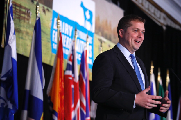 Ein Mann im Anzug und Krawatte steht an einem Rednerpult mit Mikrofon, umgeben von Flaggen und einem Bildschirm im Hintergrund, und scheint eine Äußerung über die Zahlung von 1,5 Milliarden Dollar durch die Regierung an die Milchindustrie zu machen.