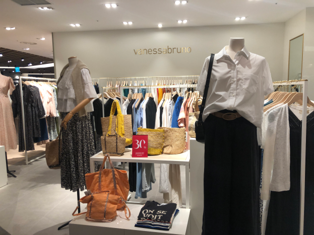 Ein Vanessa Bruno Geschäft in Singapur mit Schaufensterpuppen in Kleidern, Kleidung an Bügeln, Taschen und anderen Gegenständen auf Tischen, einer Texttafel an der Wand, einem Spiegel auf der rechten Seite und Deckenbeleuchtung.