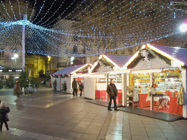 Ein lebendiger Weihnachtsmarkt in der Nacht in einer Stadt, mit Menschen, die stehen, sitzen und einer Person, die ein Kind hält, umgeben von festlichen Lichtern, Pfählen, Pflanzen, Bäumen und Gebäuden mit Fenstern unter einem bewölkten Himmel.