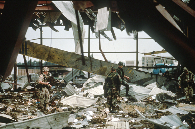 Eine Gruppe von Soldaten in Helmen und mit Gewehren durchquert die Trümmern eines zerstörten Gebäudes, mit verstreuter Trümmern, entfernten Gebäuden, Bäumen und einem klaren Himmel im Hintergrund.