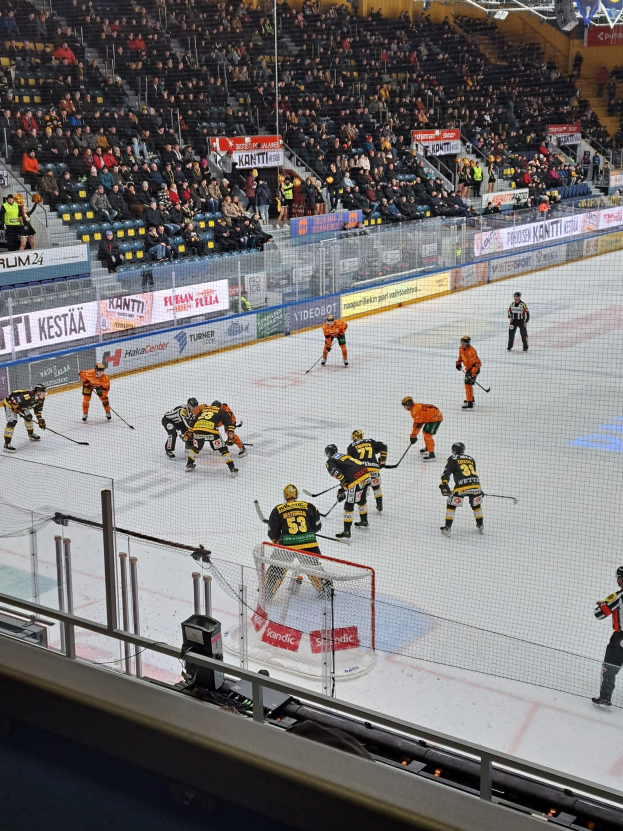 Eine Gruppe von Menschen, die Hockey auf einem Eisring spielen, mit einem Netz in der Mitte und einem Zaun drumherum, Zuschauern im Hintergrund und Bannern mit Text, unter hellen Stadionlichtern.