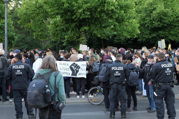 Eine große Gruppe von Menschen steht an der Straße, einige halten Schilder, andere tragen Mützen und Taschen, mit einem Fahrrad im Vordergrund und Bäumen und einem Pfahl im Hintergrund, bei einer Black Lives Matter Demonstration in Berlin.